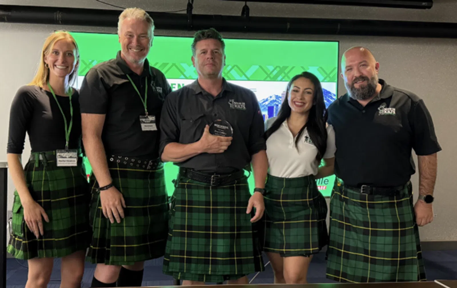 Five people wearing green plaid kilts posing together indoors at an event with a green screen behind them.