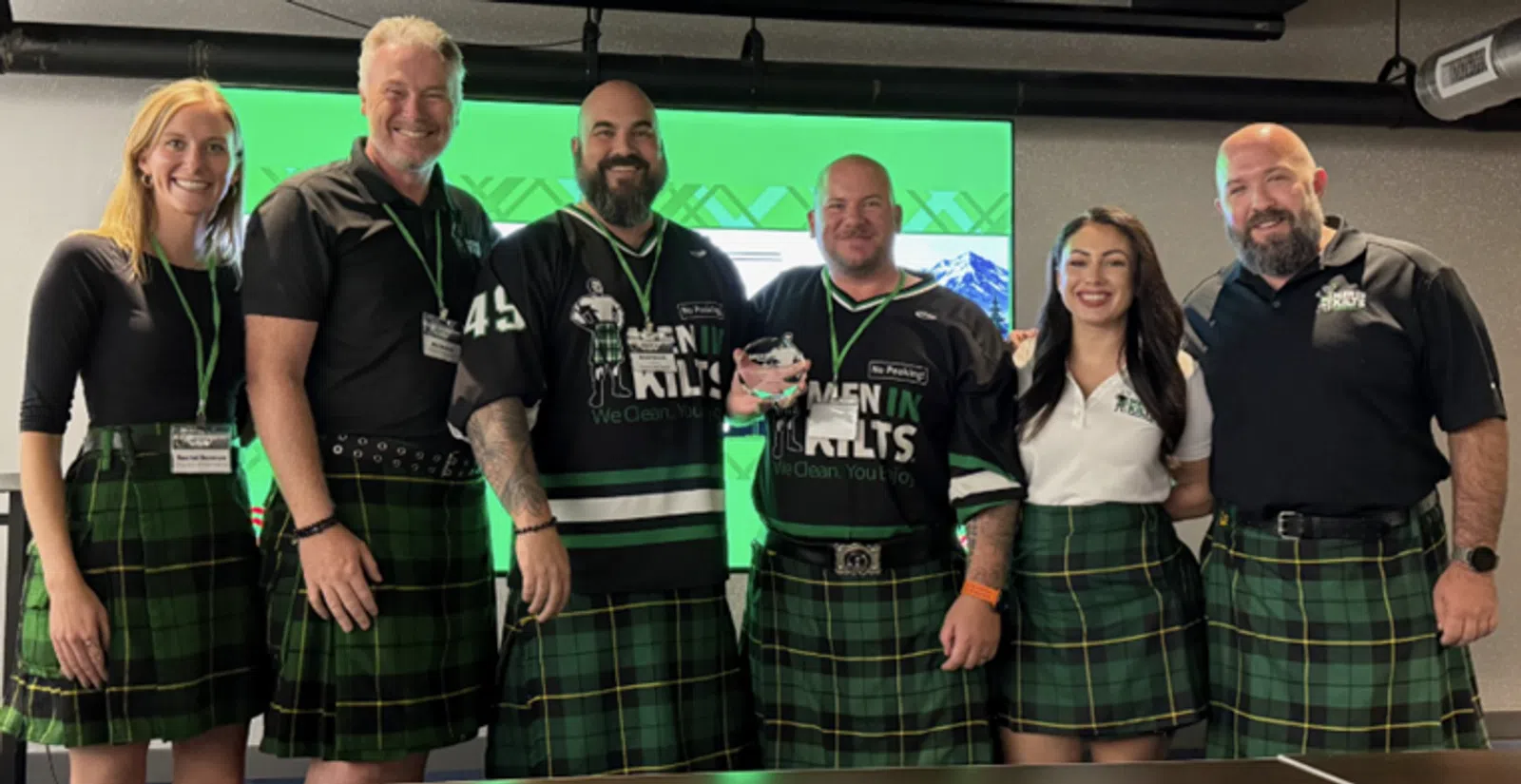 Group of six people wearing green plaid kilts and black shirts, smiling in front of a green screen backdrop.