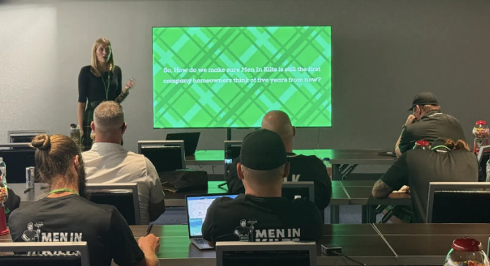 Presenter giving a presentation to seated attendees with a green slide about Men In Kilts company future vision.