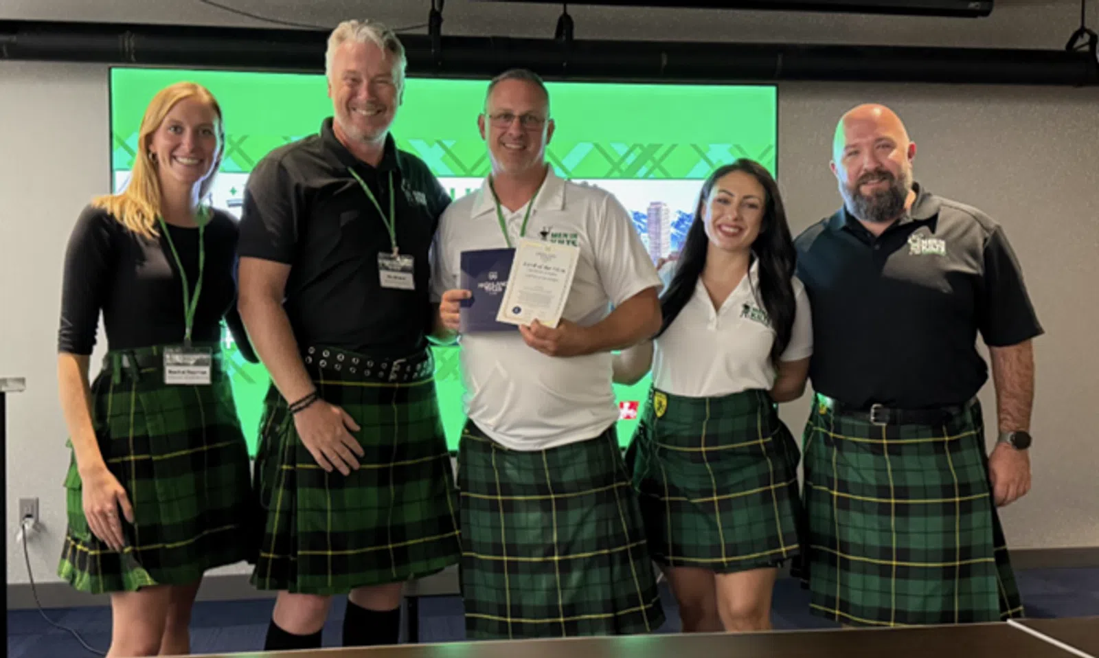 Group of five people wearing green plaid kilts, smiling and holding certificates in front of a green screen.