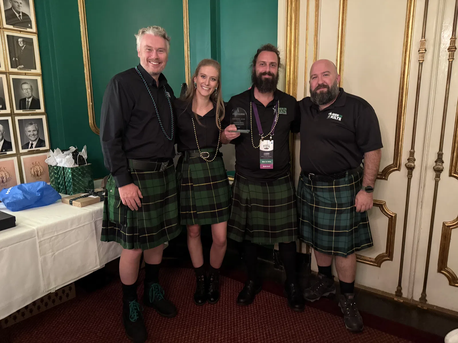 Four people wearing green plaid kilts posing indoors, smiling and holding an award, standing in ornate room.
