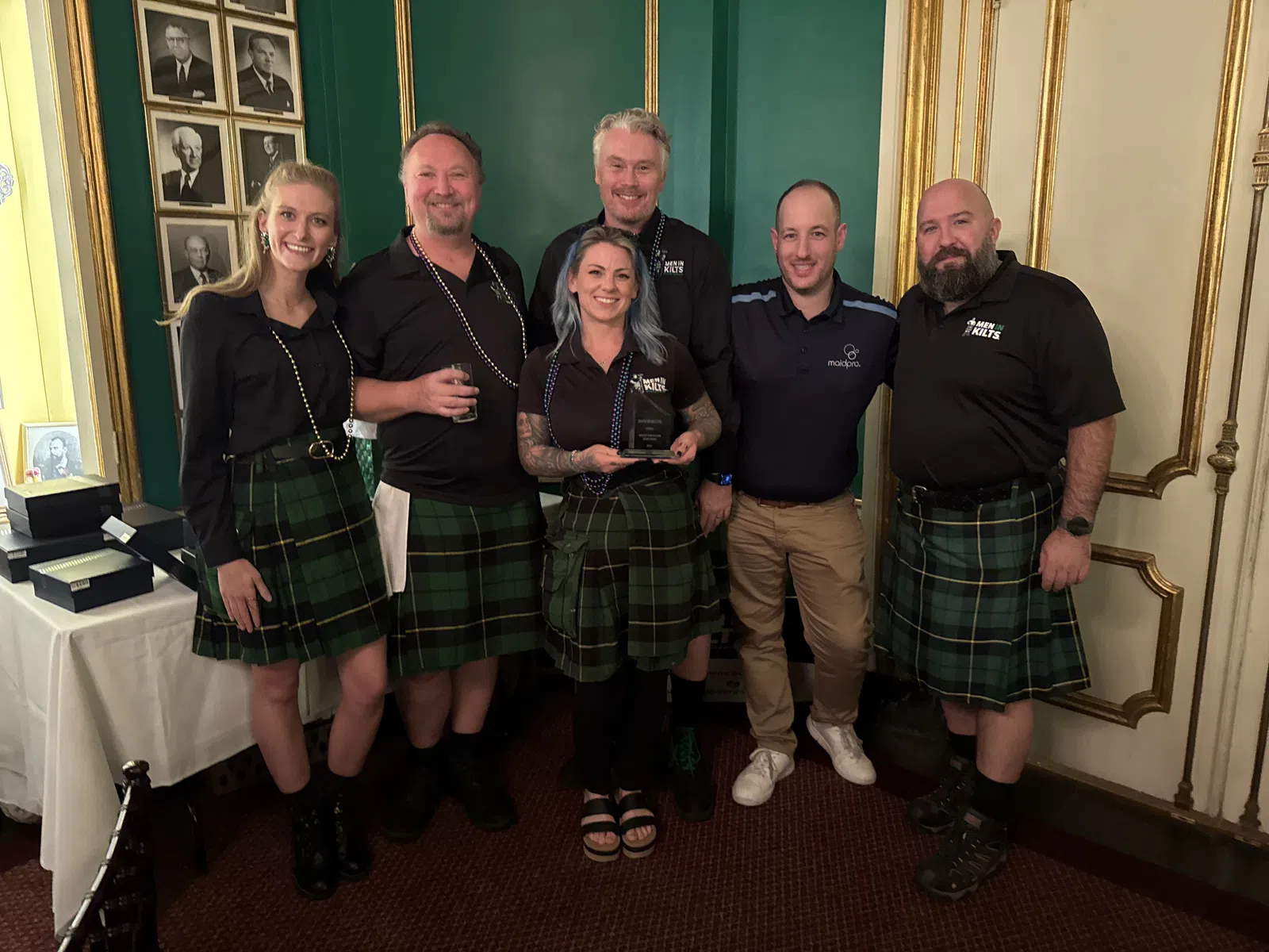 Group of six people, five wearing green tartan kilts, posing indoors with award in decorated room.