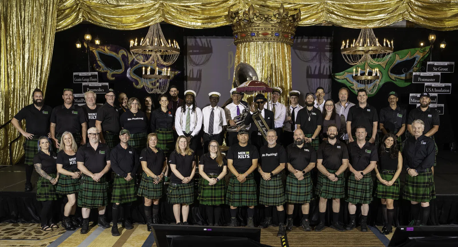 Group photo of men and women wearing green plaid kilts and black shirts posing on stage under gold curtains and chandeliers.
