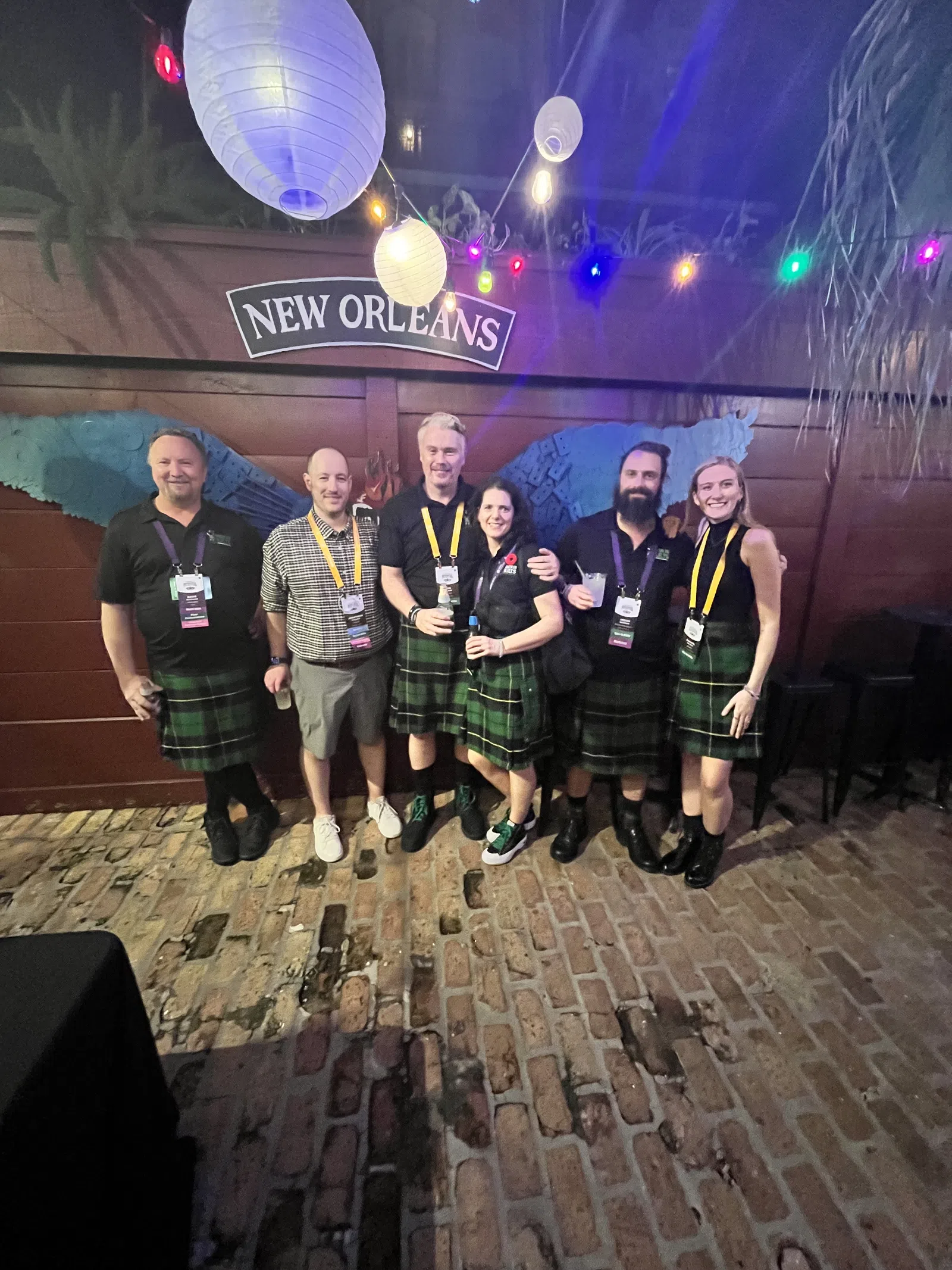Group of six people wearing kilts at a New Orleans venue with hanging lanterns and colorful string lights