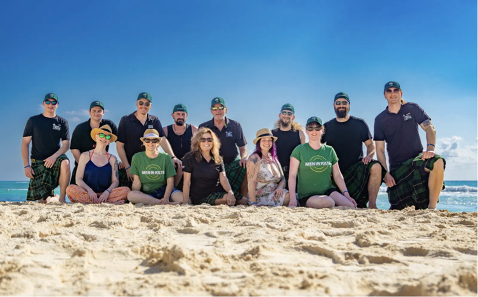 Group of people in kilts and casual wear posing on sunny beach with blue sky and ocean in background
