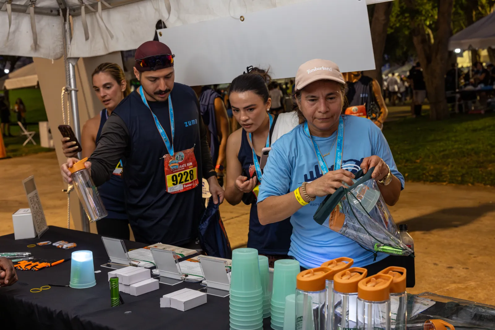 People browsing promotional water bottles and merchandise at an outdoor event booth under a tent.