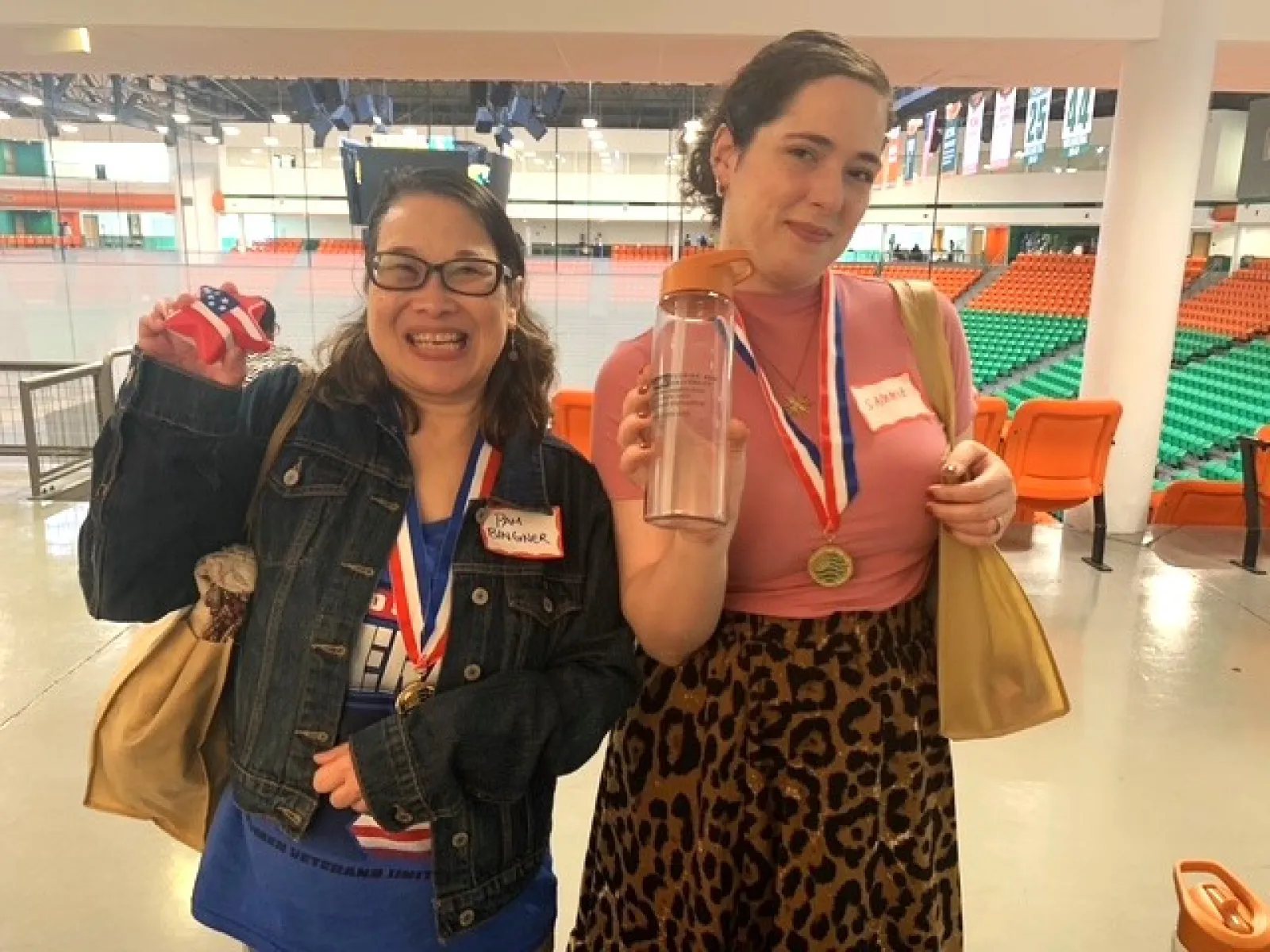 Two women wearing medals smile and hold trophies inside an indoor sports arena with orange and green seats.