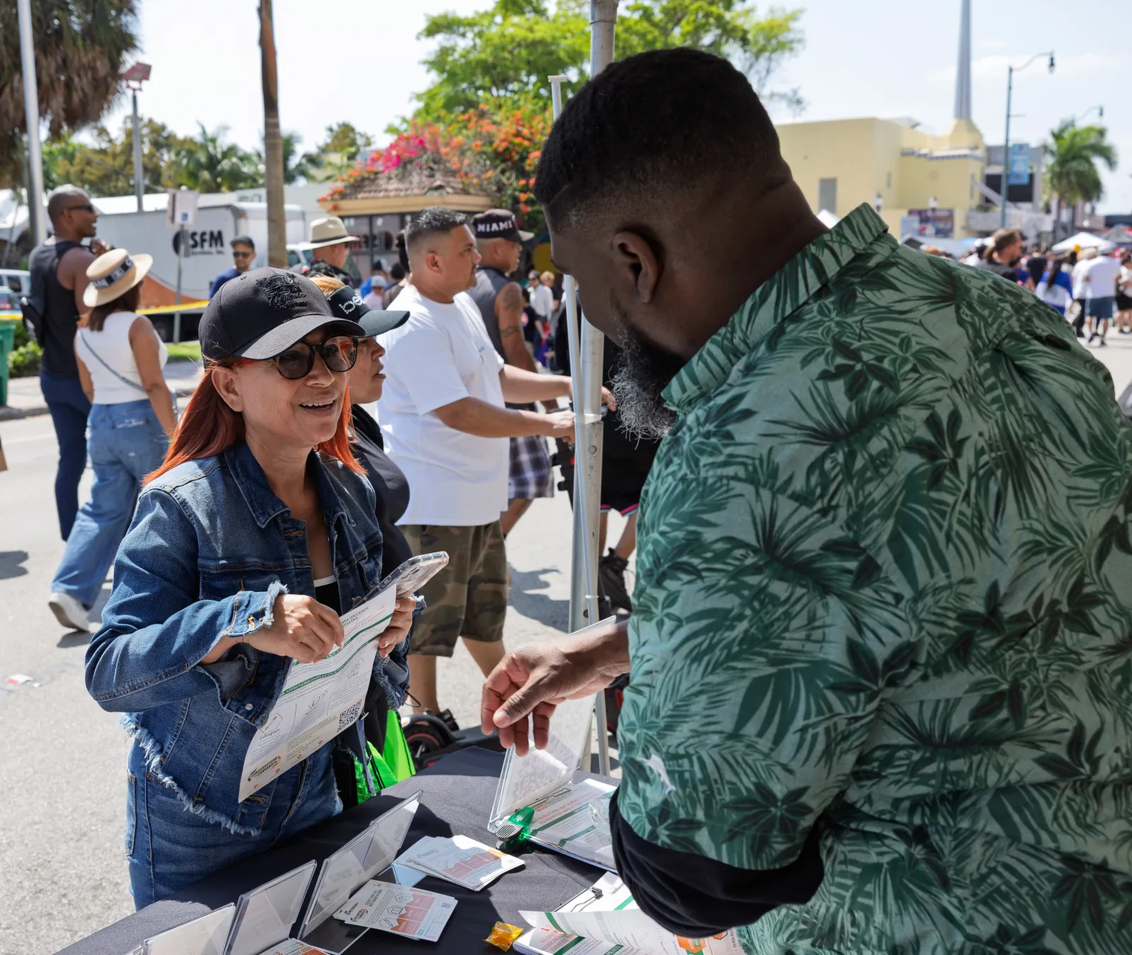 Woman in denim jacket and cap engaging with man in green shirt at outdoor event booth on sunny day