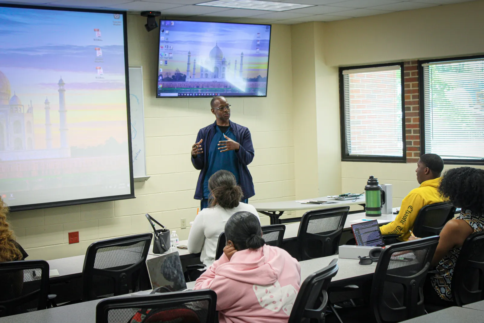 Instructor giving a classroom lecture to diverse students using projected screen and monitor displays.