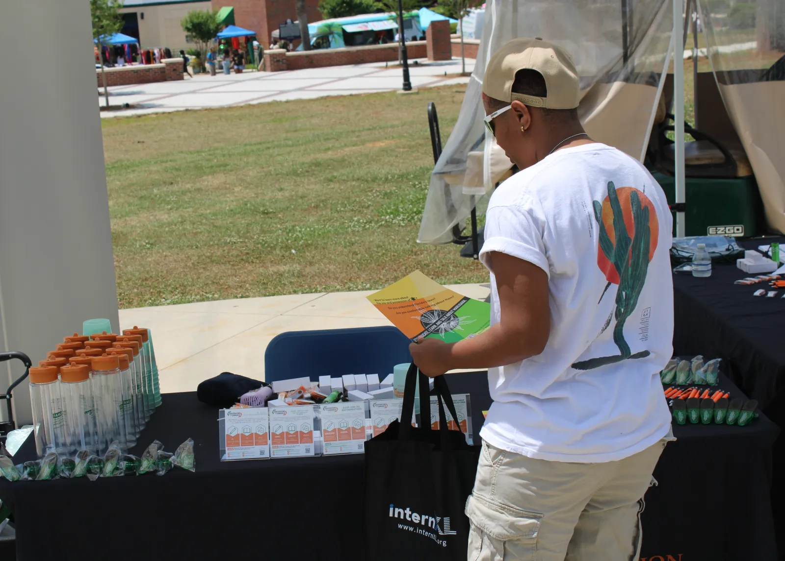 Person browsing an outdoor vendor table with promotional items and brochures on a sunny day.
