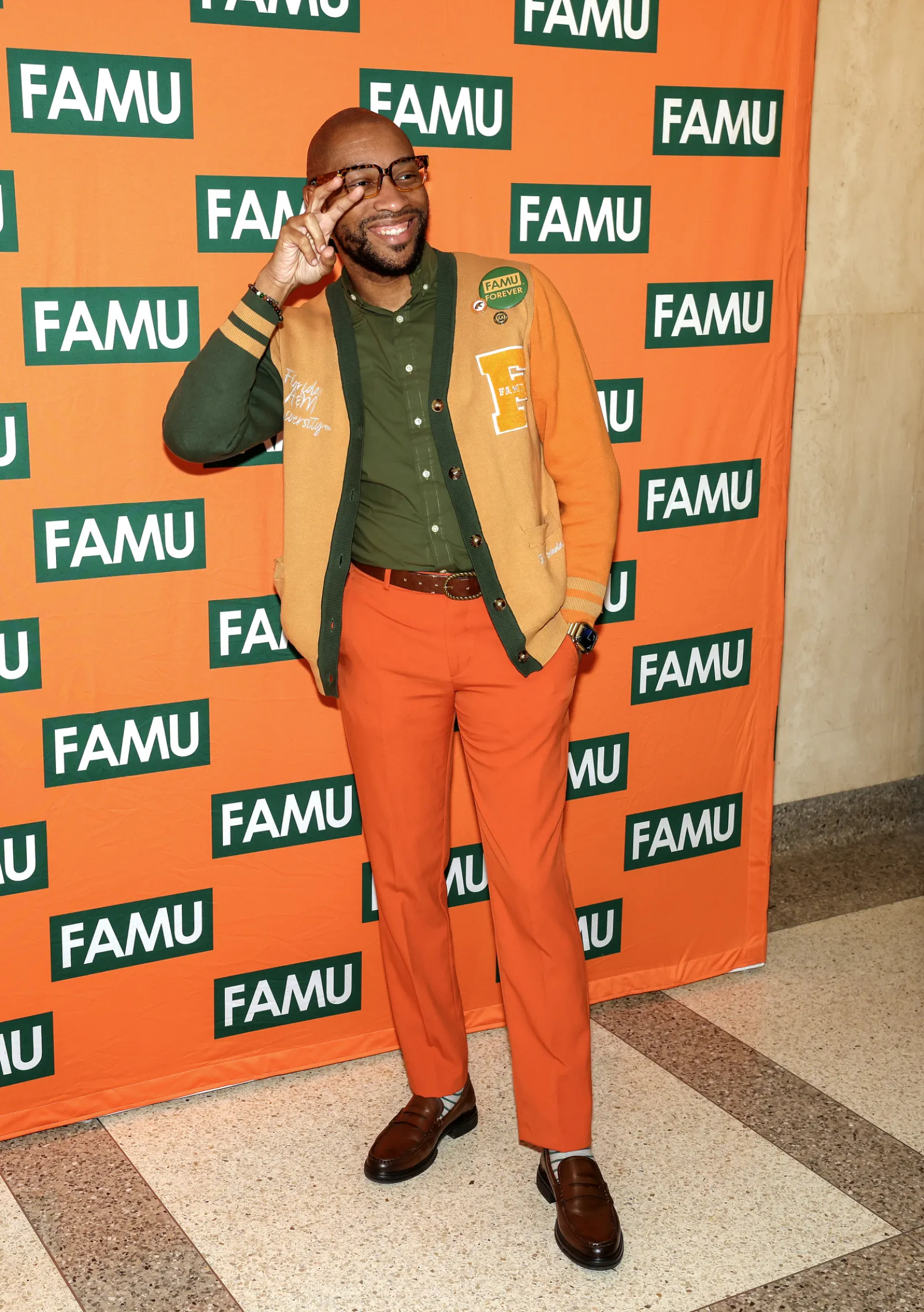 Man in orange and green outfit posing joyfully with FAMU backdrop at an indoor event