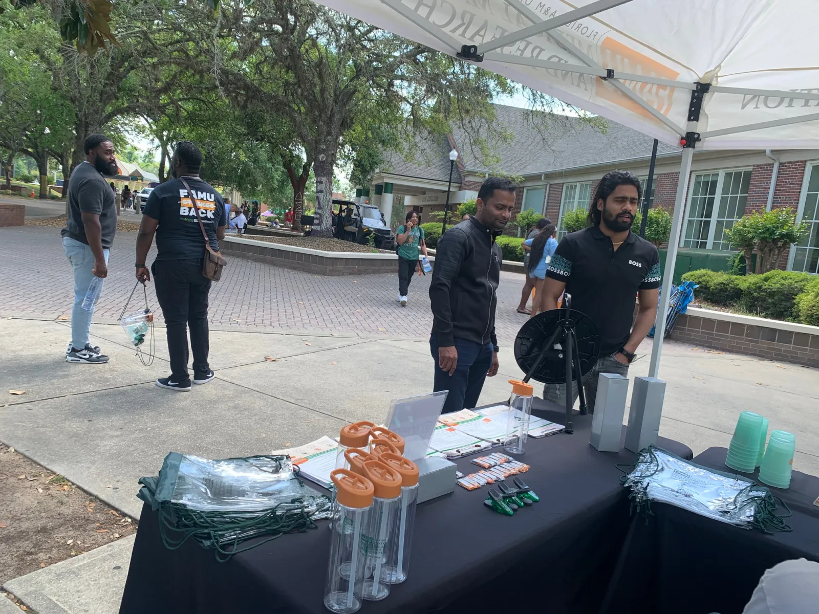 Outdoor campus event with information booth offering promotional items and two men staffing the table under a canopy.