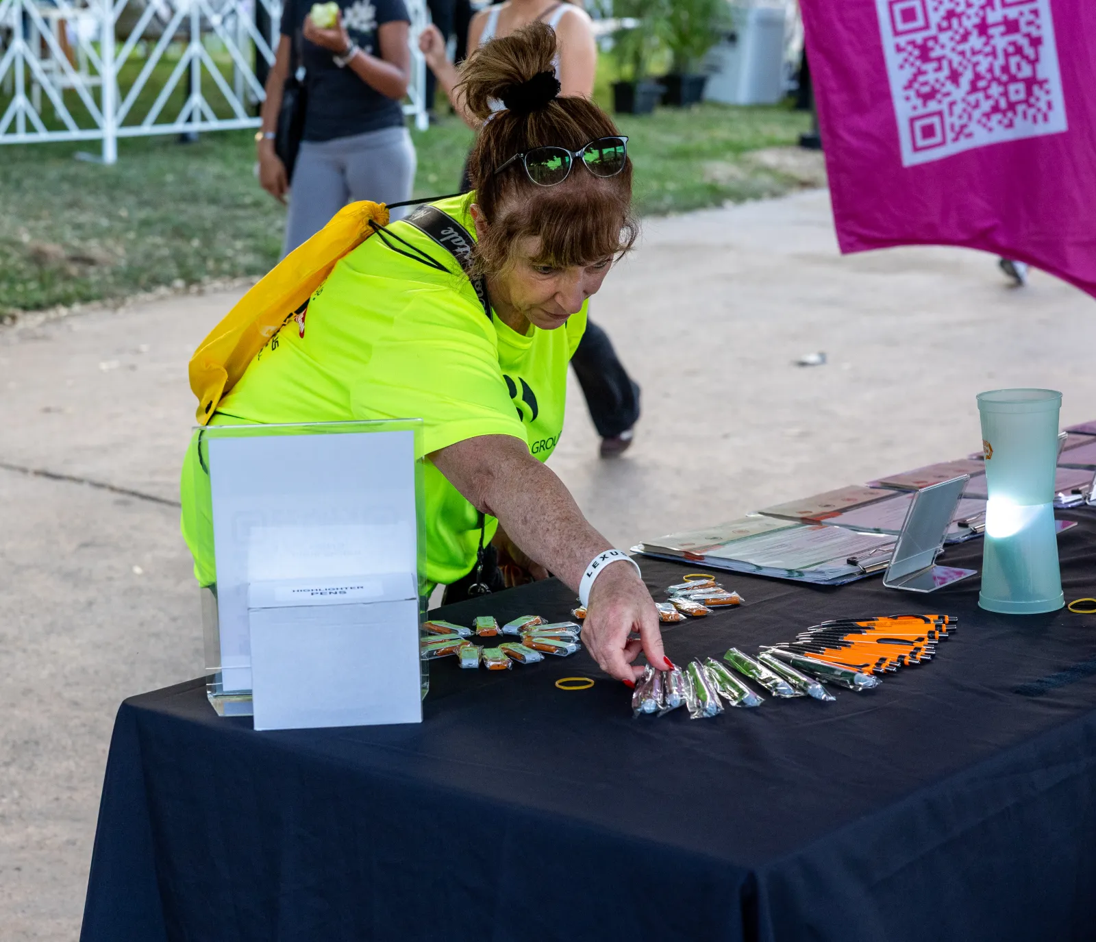 Woman in neon yellow shirt arranging promotional items on a black table at an outdoor event.
