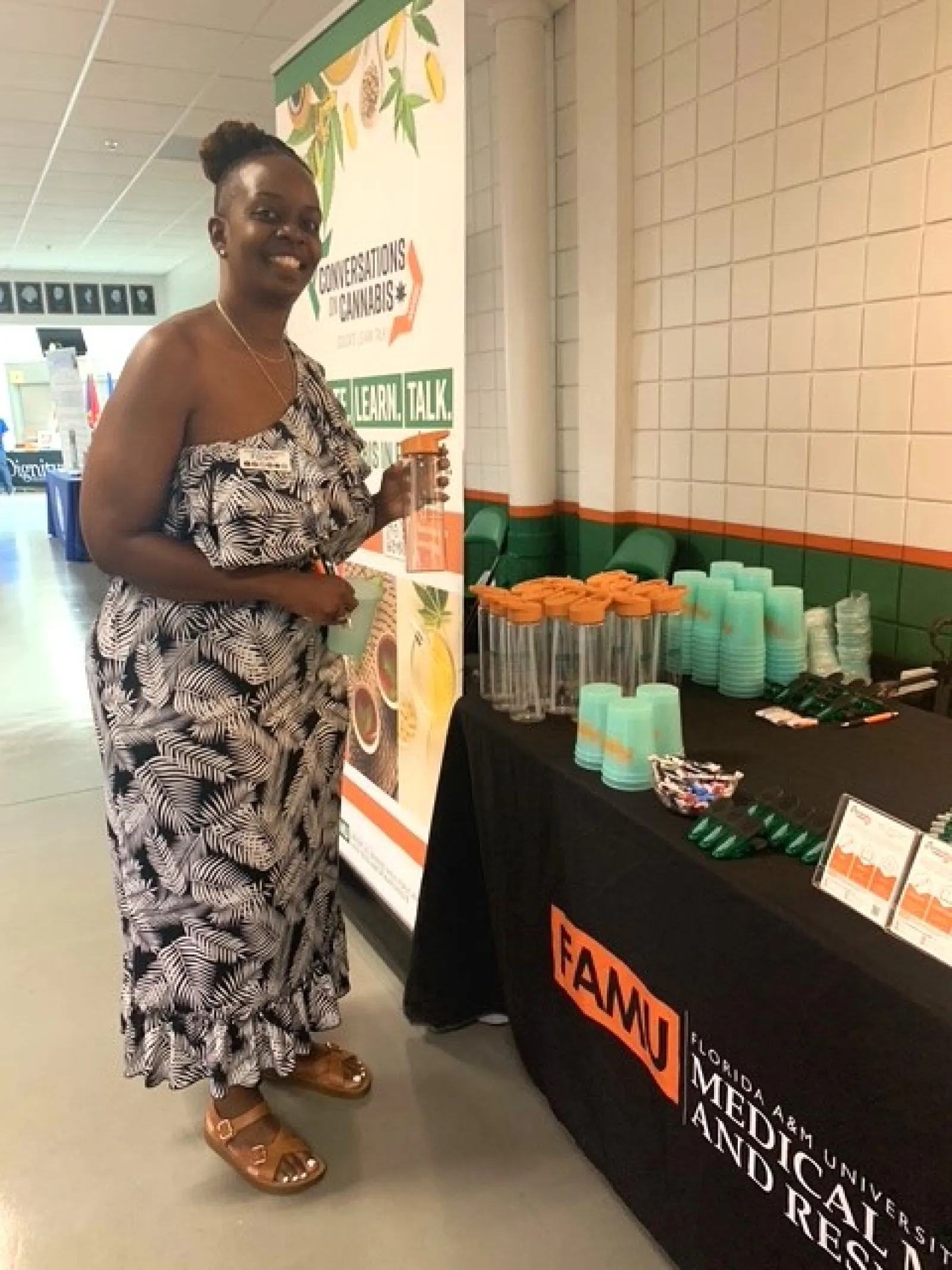 Woman in patterned dress stands by FAMU table with promotional cups and educational cannabis awareness banner.