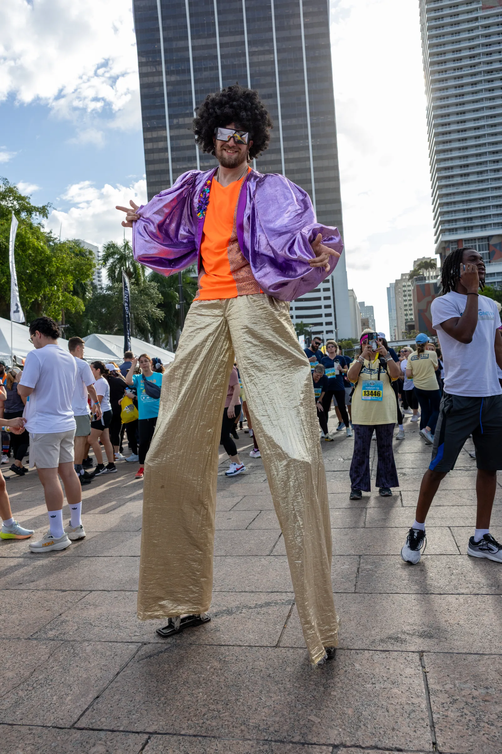 Man on stilts wearing shiny purple jacket, orange shirt, gold pants, and afro wig at outdoor city event.
