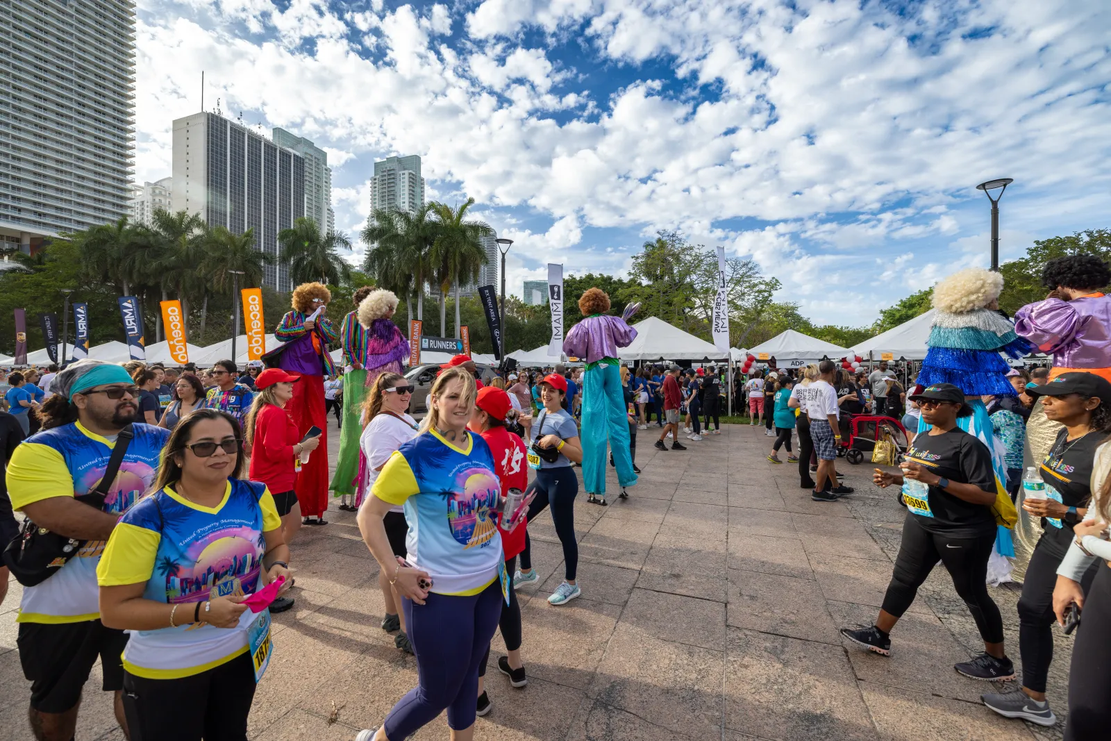 Crowd at outdoor event with performers on stilts, colorful costumes, tents, and modern buildings under a partly cloudy sky.