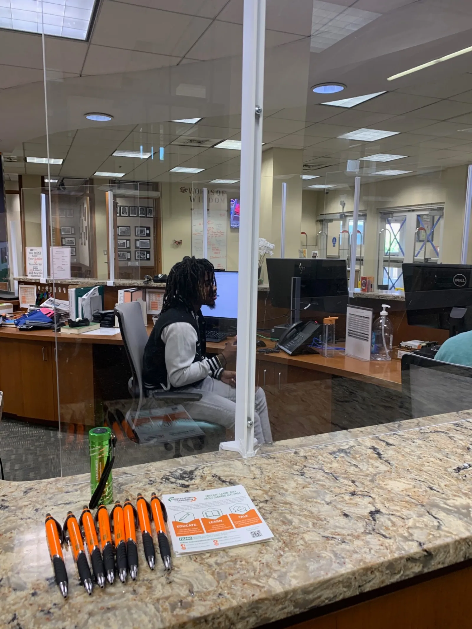 Office reception desk with pens, a water bottle, acrylic divider, and a person working on a computer.