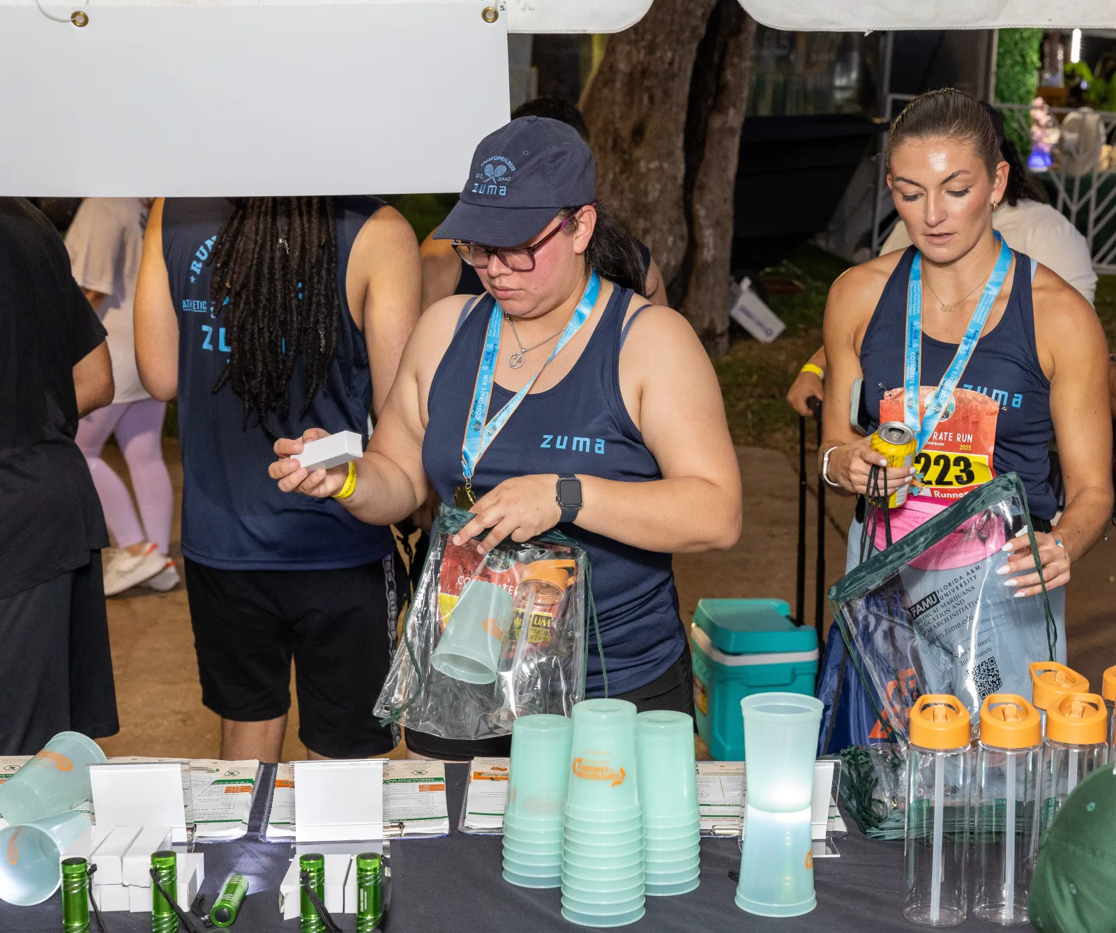 Two women wearing Zuma shirts distributing cups and water bottles at an outdoor event booth.
