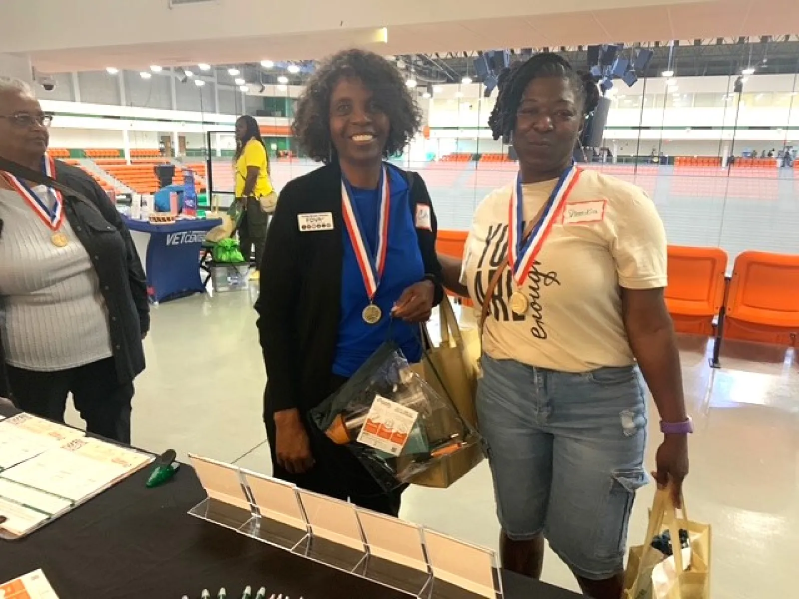 Two women wearing medals holding gift bags at an indoor event with orange seating in the background