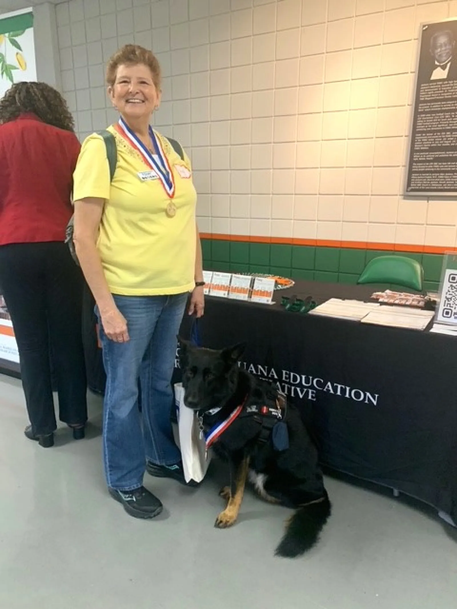 Woman in yellow shirt standing next to a German Shepherd service dog at an education event booth.