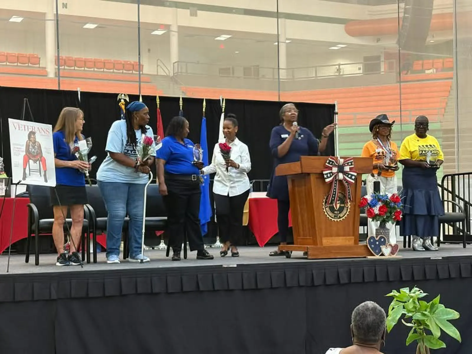 Group of women on stage holding flowers and awards during a veterans appreciation event in a gymnasium.