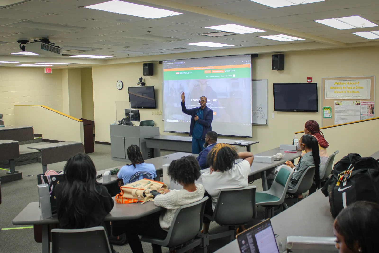 Instructor giving a lecture to students in a brightly lit classroom with projection screen and desks.