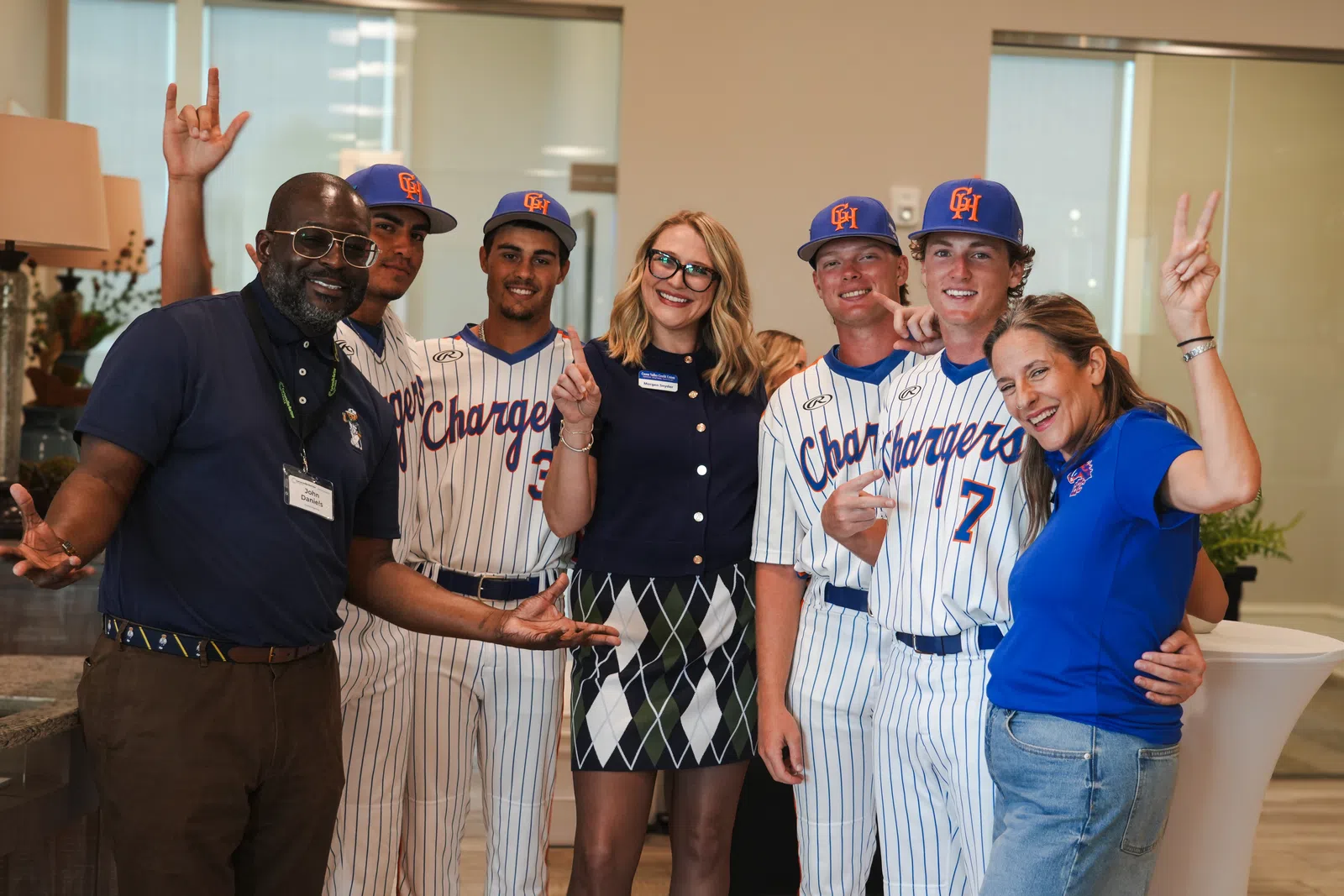 Group of happy people including baseball players and staff posing indoors with smiles and friendly gestures.