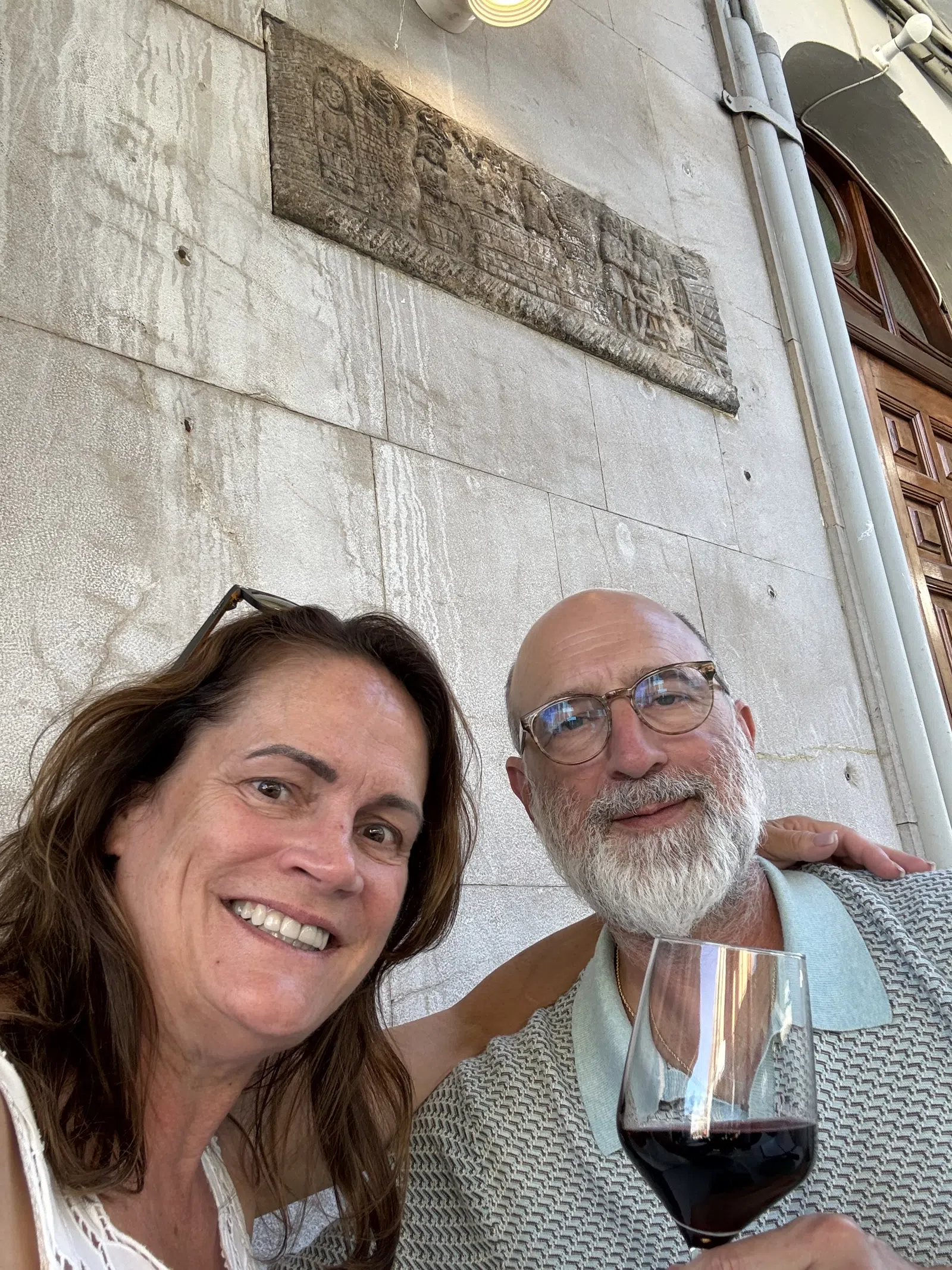 Smiling couple enjoying red wine at outdoor cafe with stone wall and wooden door background.