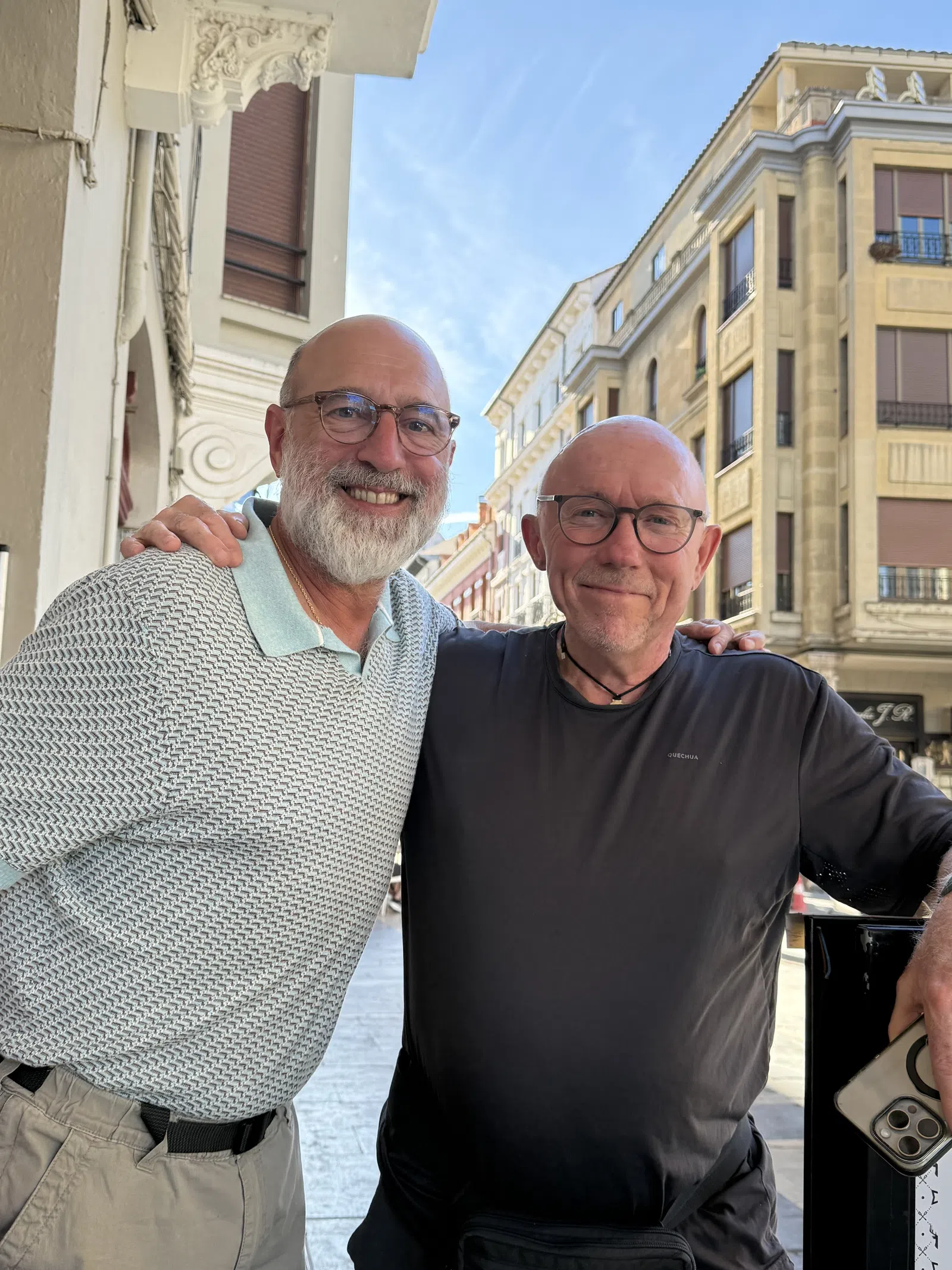 Two older men smiling and posing together on a sunny street with historic buildings in the background.