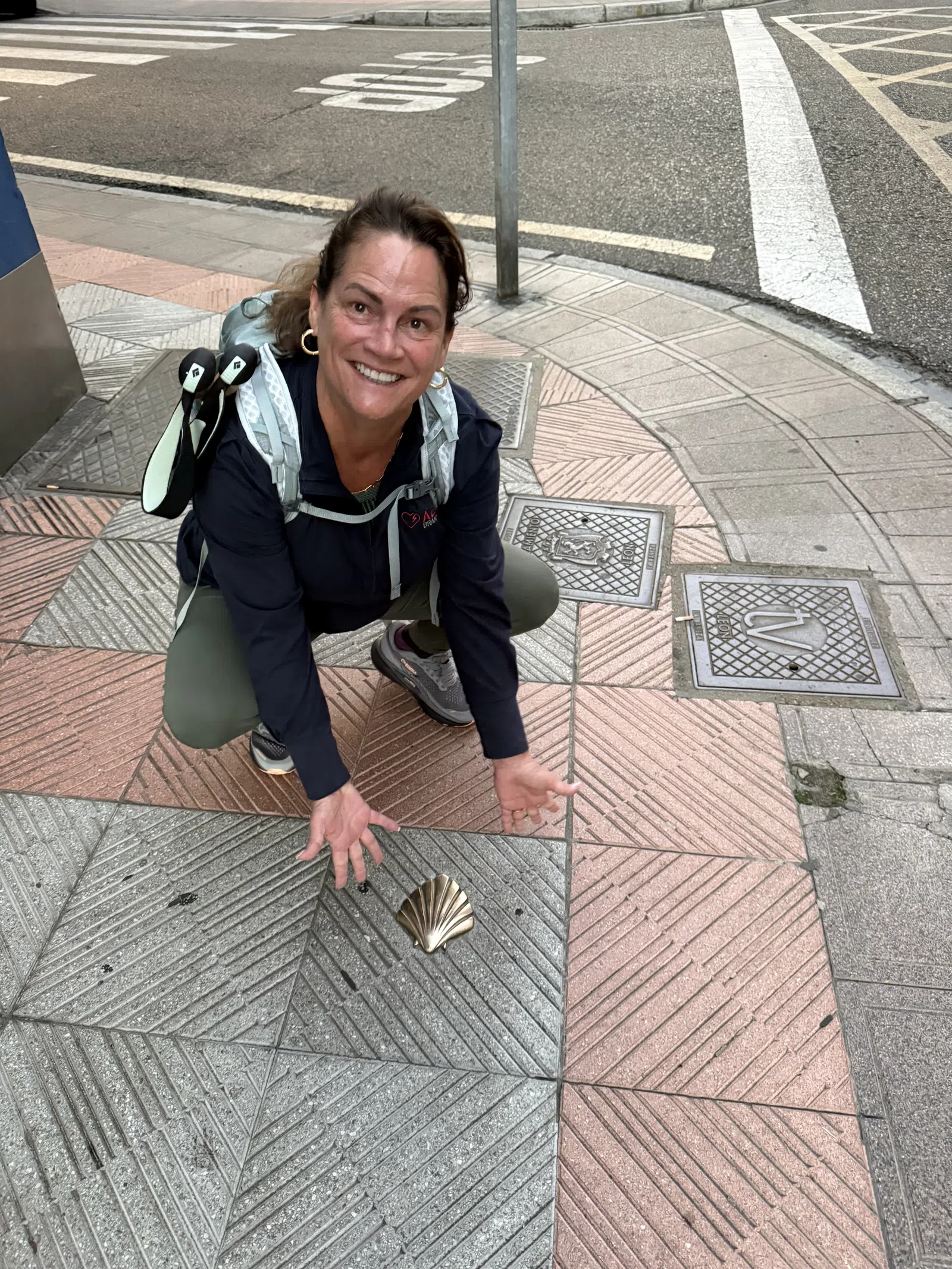 Smiling woman crouching on patterned sidewalk pointing at a bronze scallop shell embedded in the pavement.