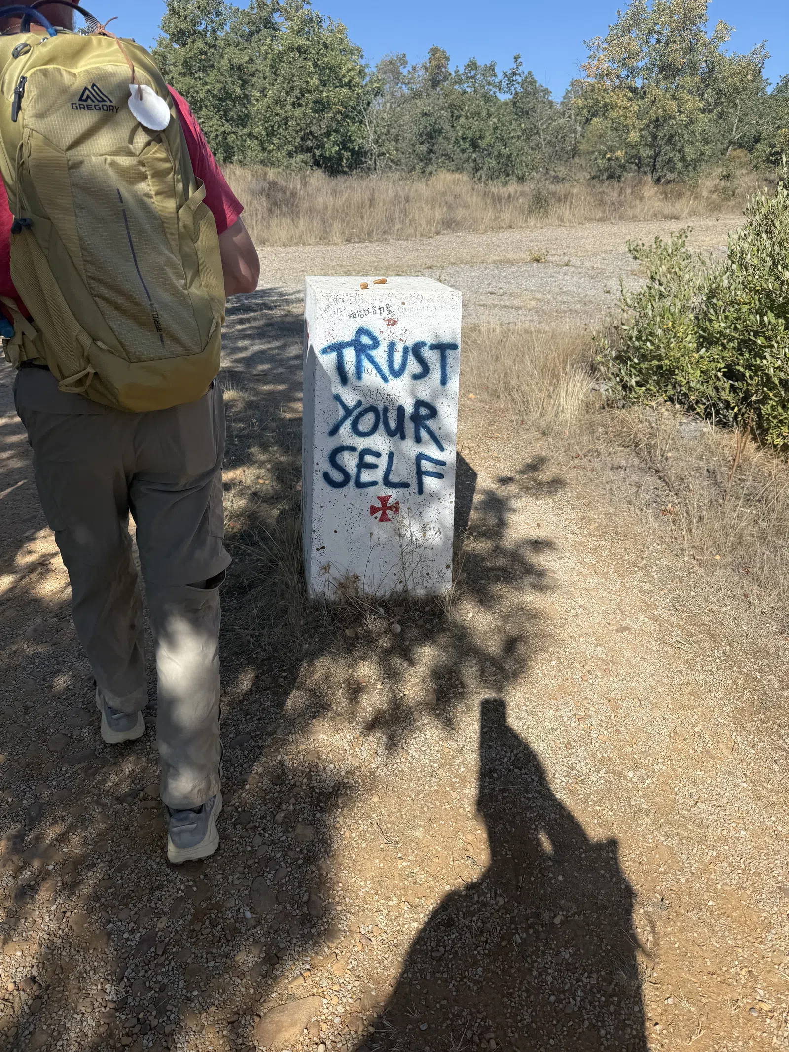 Hiker with yellow backpack walks past a concrete marker reading 'TRUST YOUR SELF' in blue graffiti on a sunny trail.