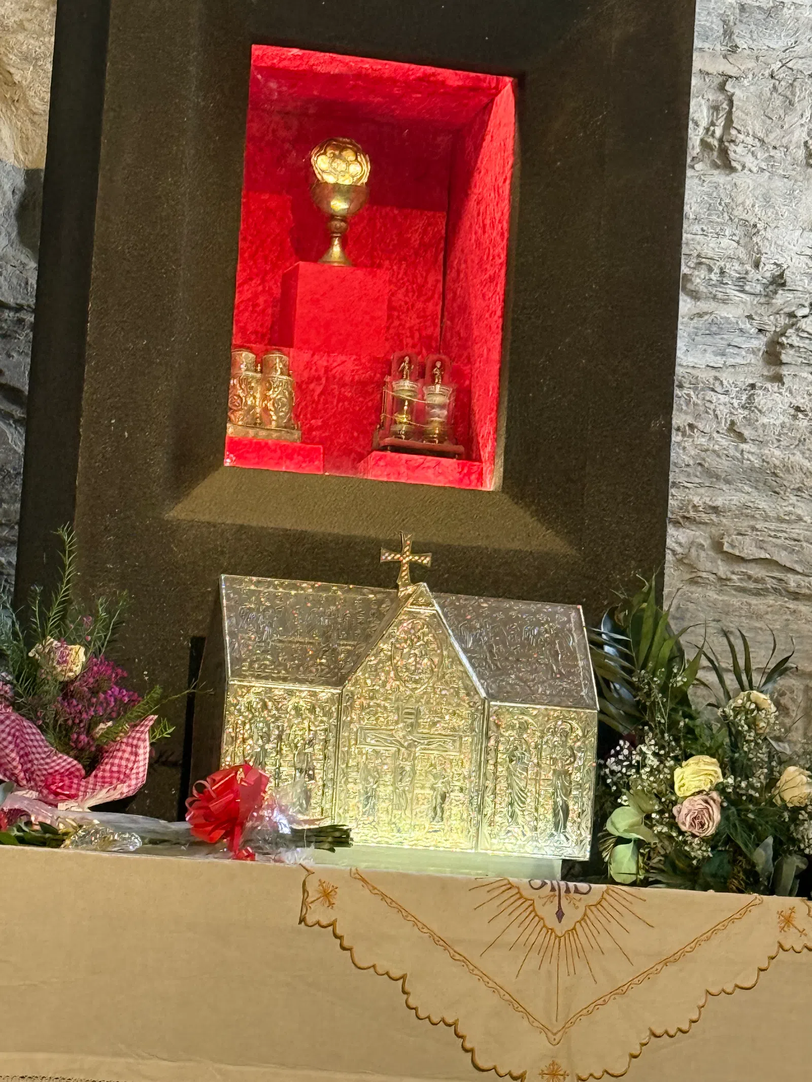 Silver religious relic shrine with intricate carvings on altar surrounded by flowers in stone chapel niche.