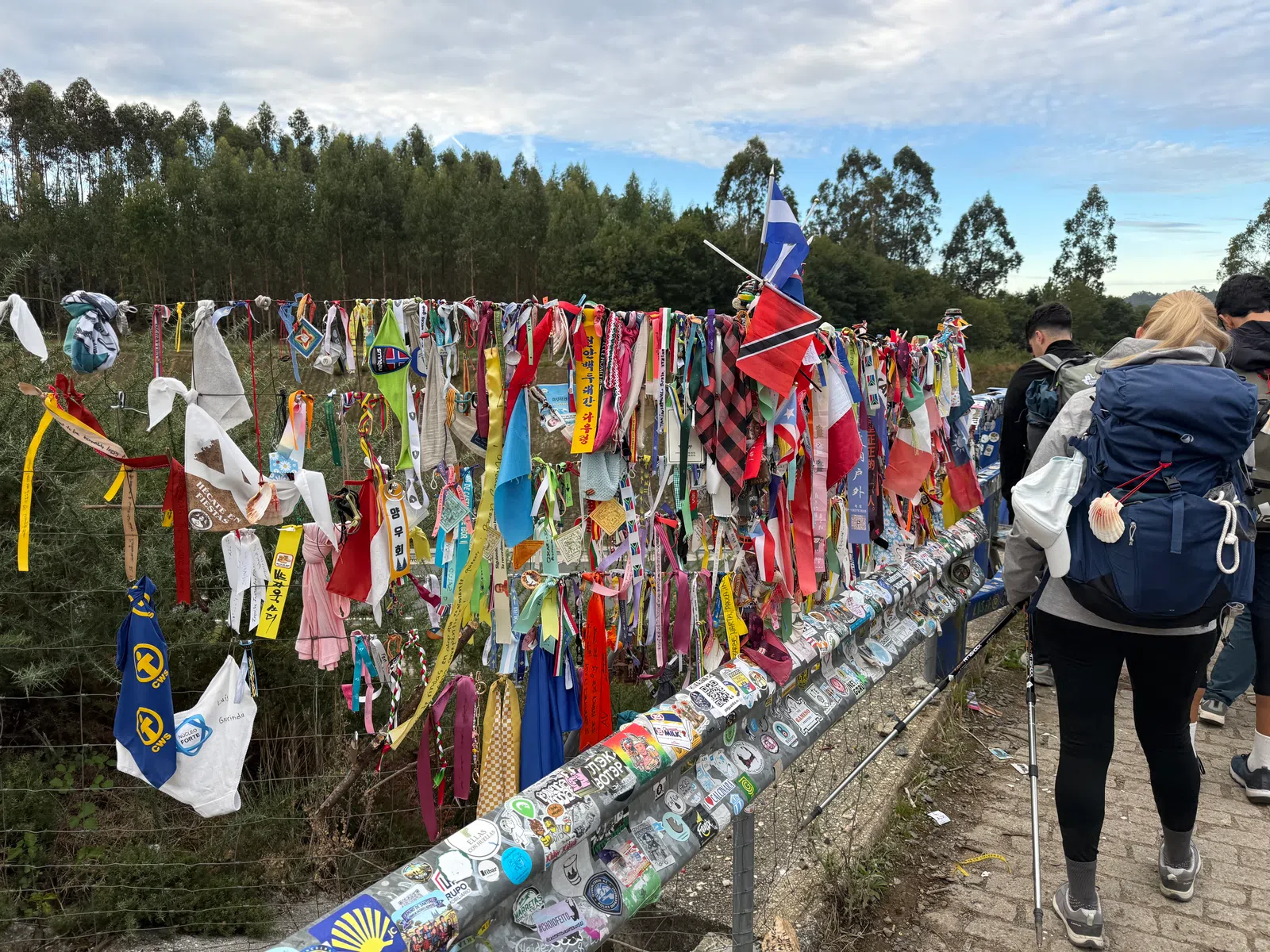 Colorful fabric strips and flags tied to wire fence along a trail with hikers walking nearby under a cloudy sky.