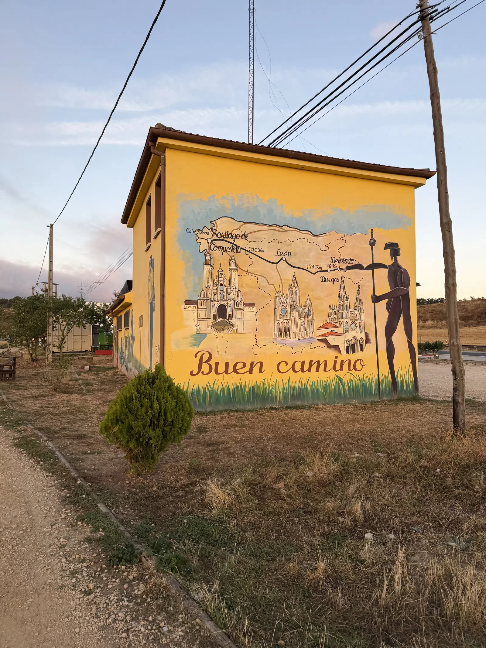 Yellow building with mural showing Camino de Santiago route map, landmarks, pilgrim figure, and Buen camino text.