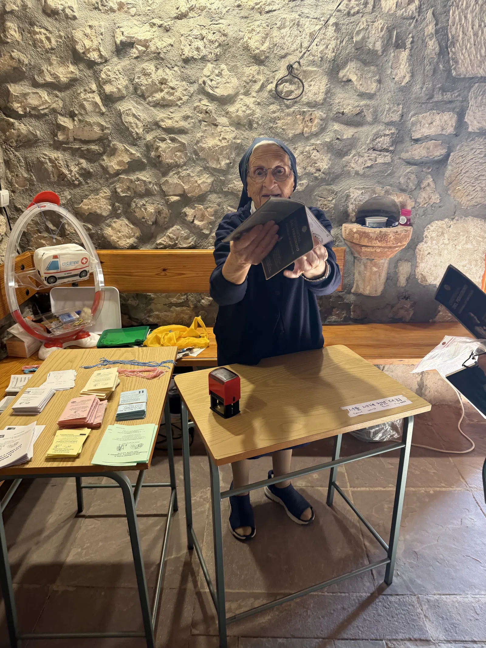 Elderly woman in headscarf handing out booklets at a rustic stone-walled room with tables of pamphlets.