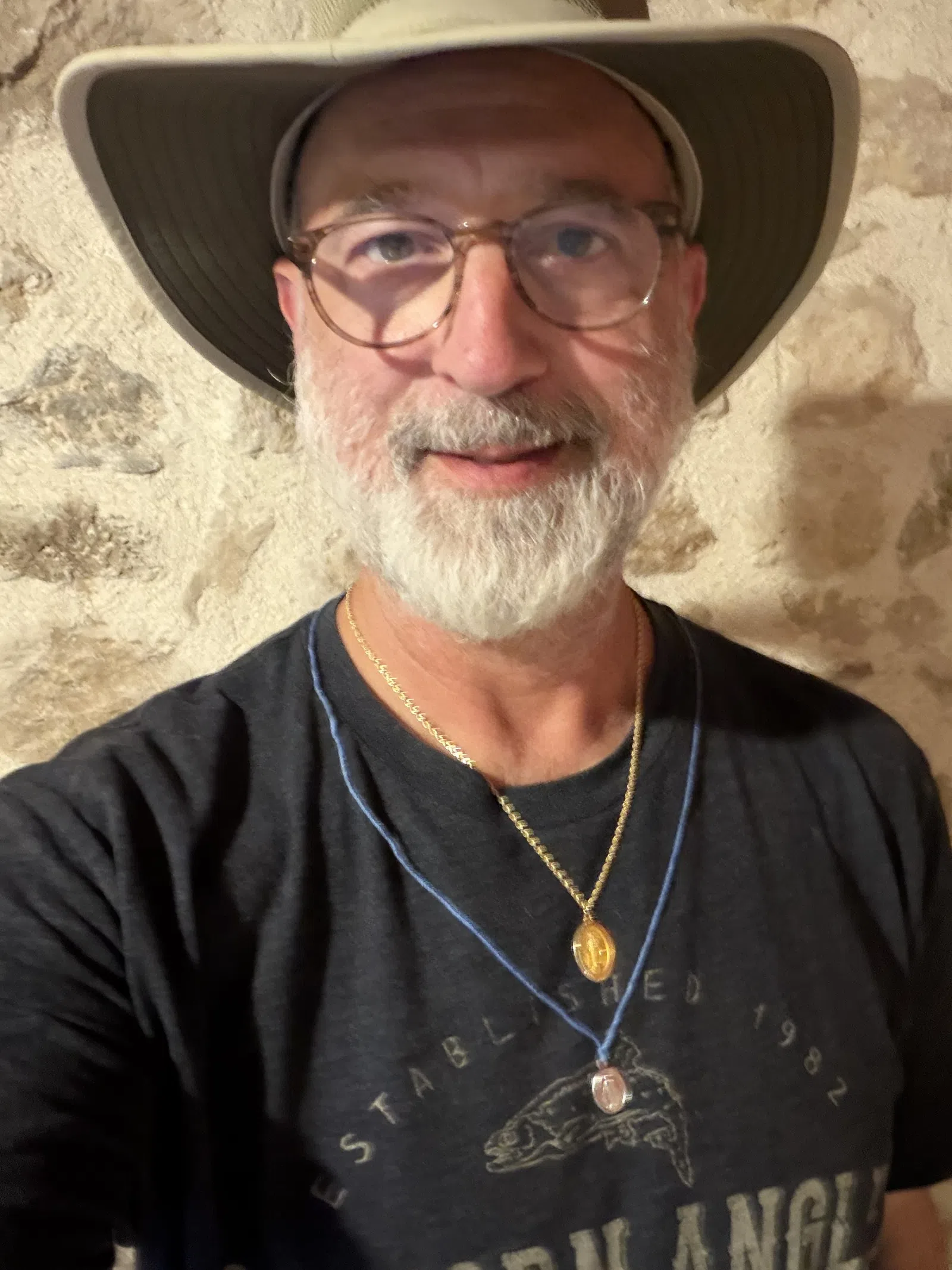 Middle-aged man with glasses and white beard wearing a wide-brim hat, two necklaces, and a black graphic t-shirt.