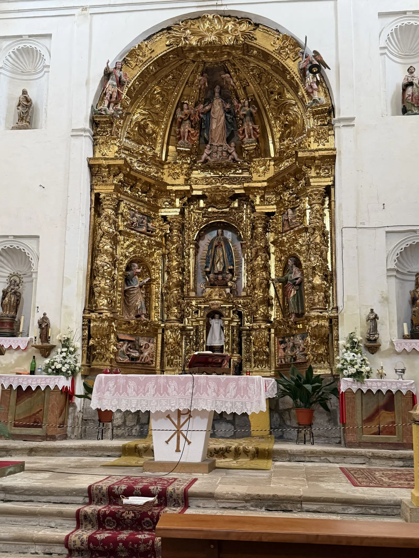 Ornate gold religious altar with statues, white and pink altar cloths, floral arrangements, and stone steps in a church.