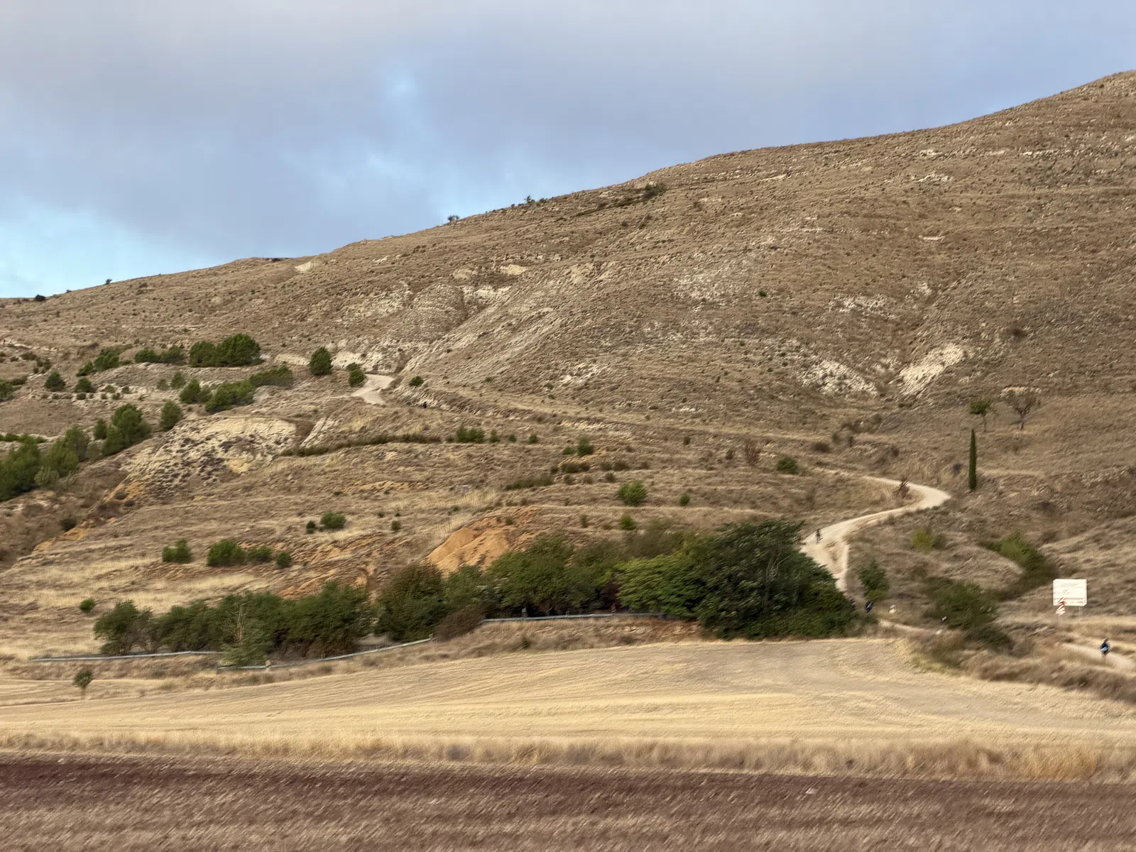 Curving dirt road winding up a dry, hilly landscape with sparse vegetation under a cloudy sky.
