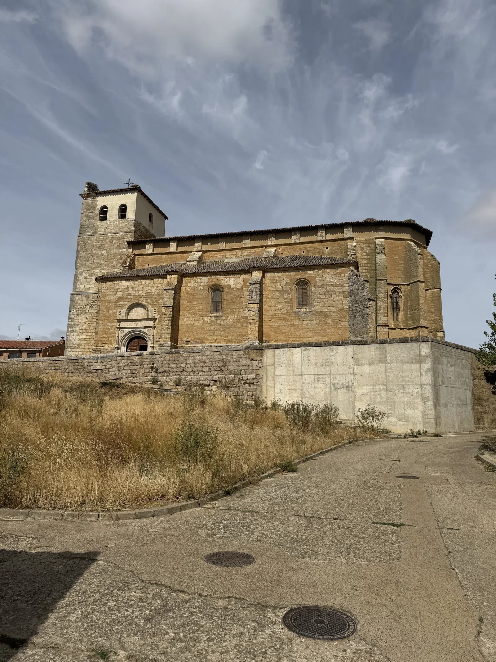 Historic stone church with bell tower under a cloudy blue sky beside a curved paved road and dry grass.