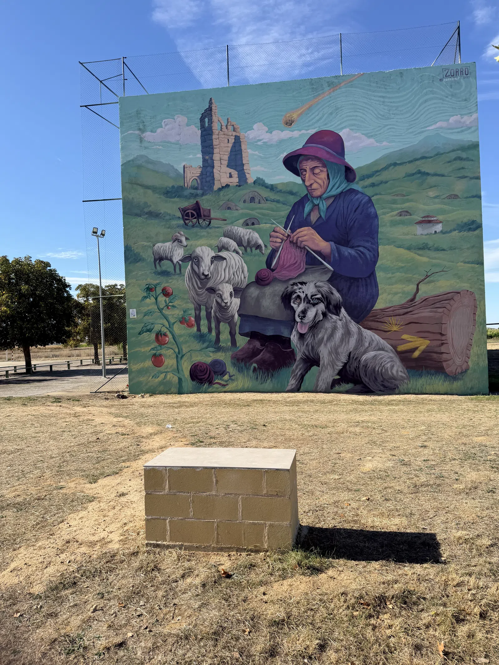 Mural of an elderly woman knitting with sheep, a dog, and rural ruins under a blue sky with a comet above.