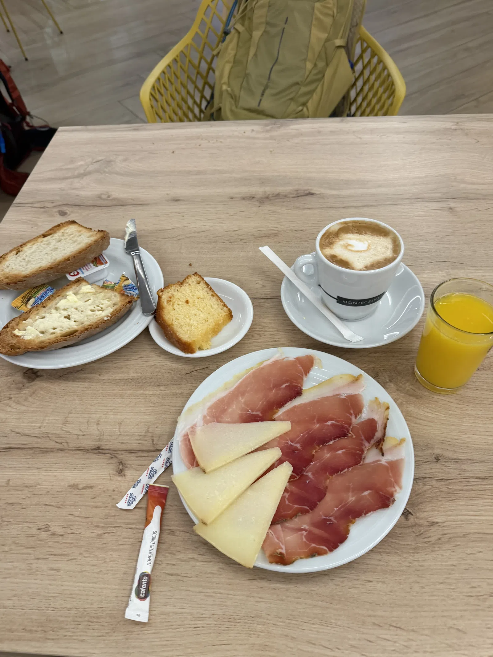 Breakfast with sliced cured ham, cheese, toasted bread, coffee with milk, orange juice, and a small cake on a wooden table.