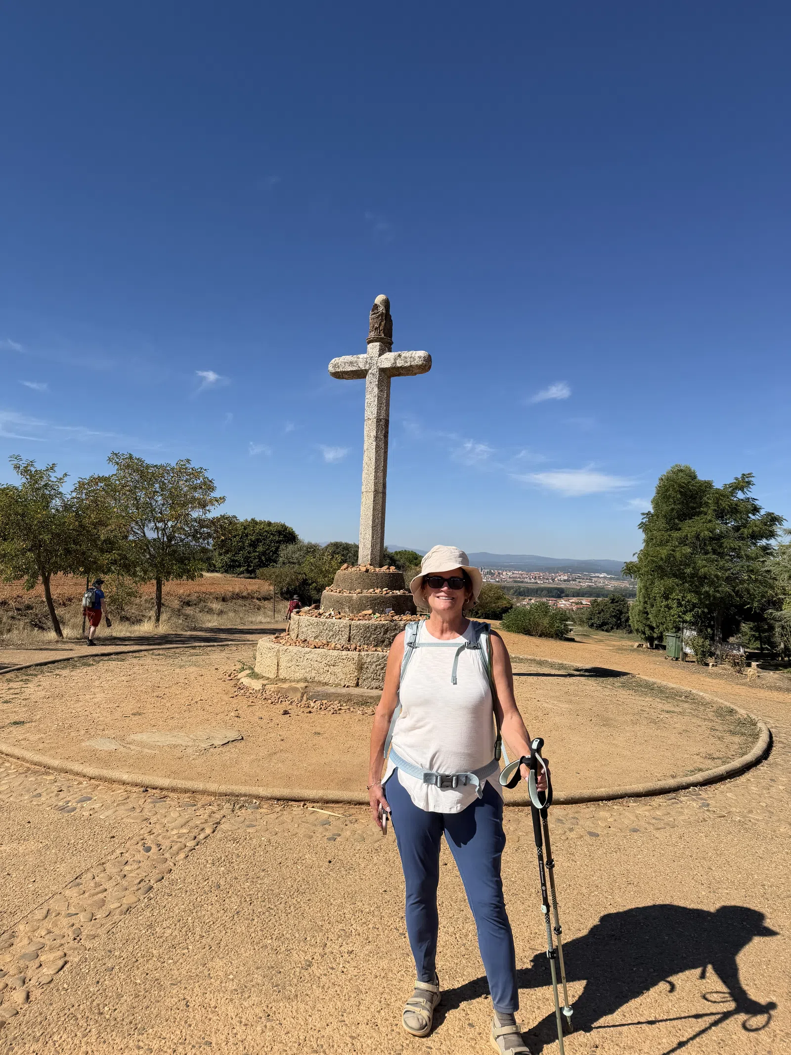 Smiling woman hiker with trekking poles standing in front of a large stone cross on a sunny day with clear blue sky.