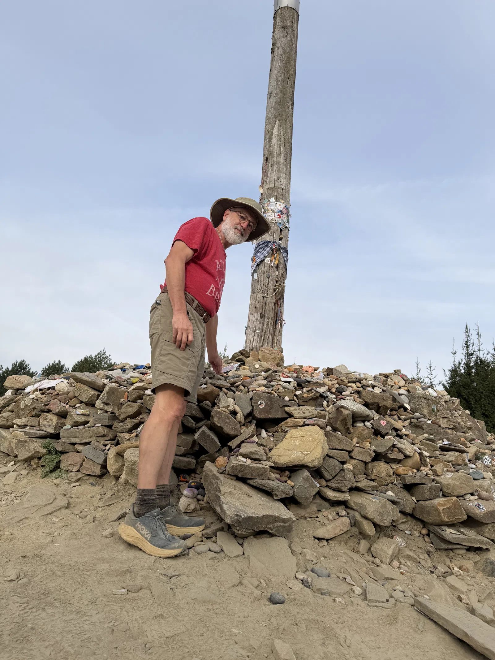 Man in red shirt and hat standing beside a tall wooden post on a large pile of rocks outdoors