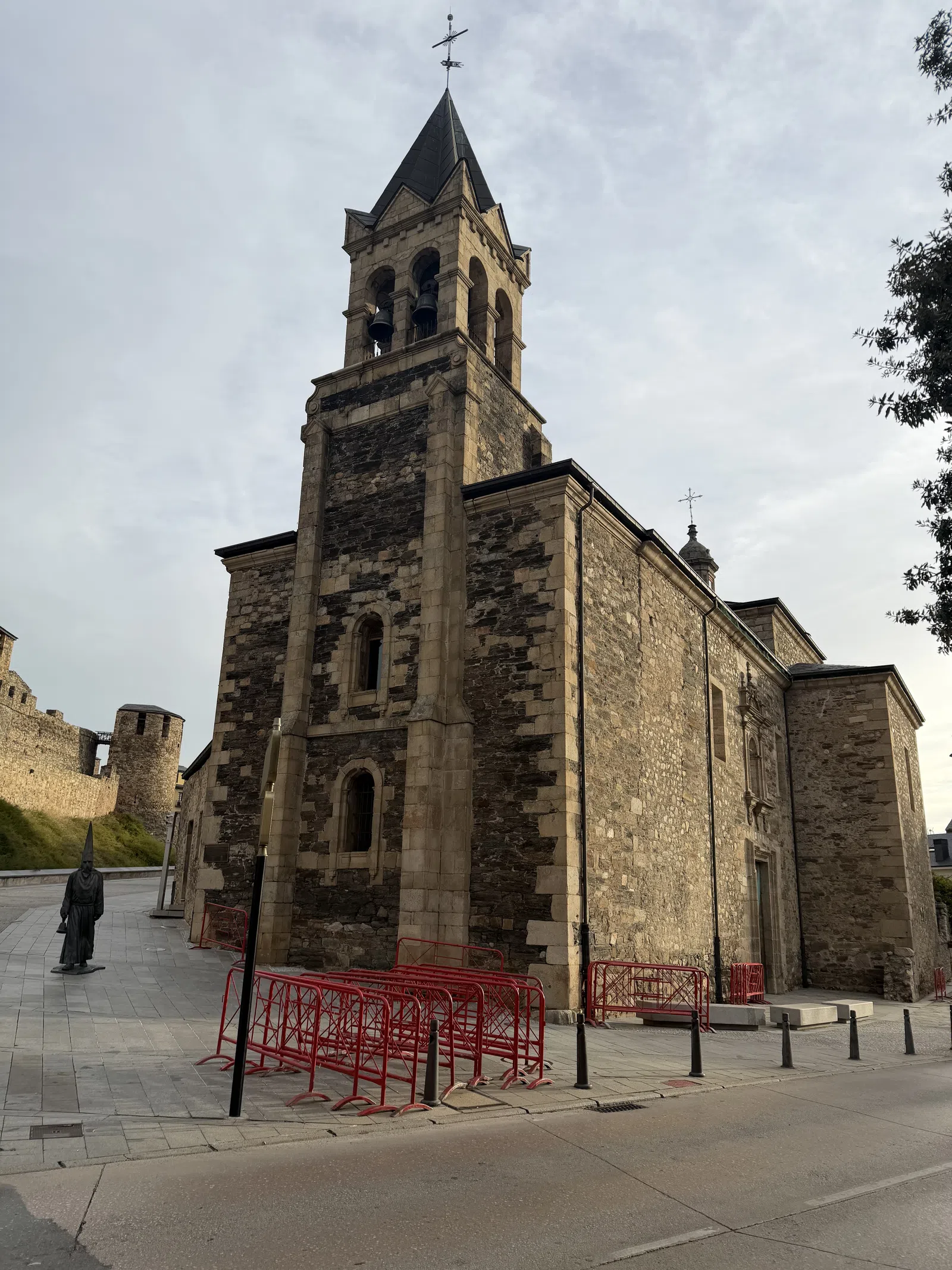 Historic stone church with bell tower and red metal barriers on sidewalk under cloudy sky.