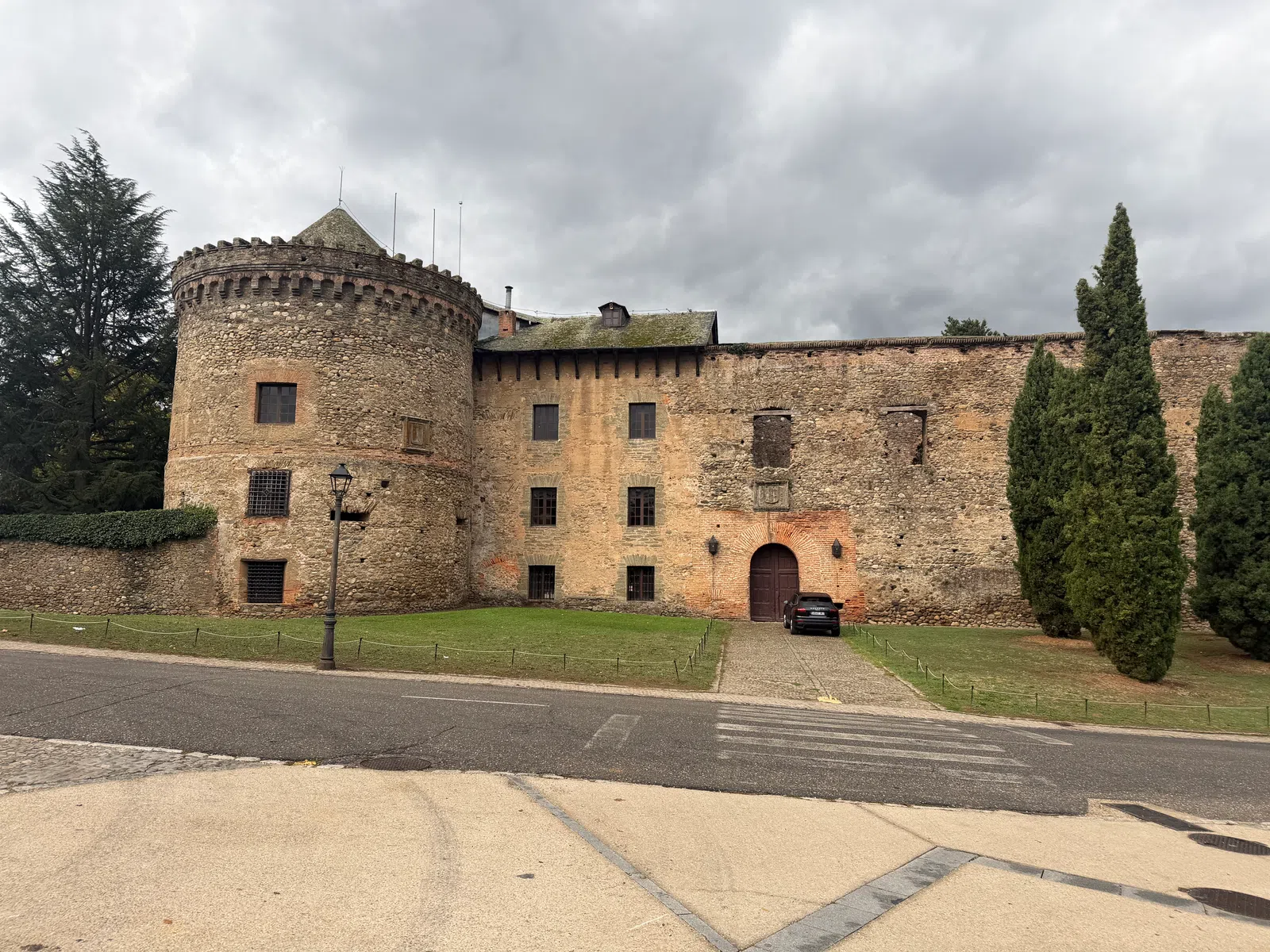 Historic stone castle with round tower and arched wooden door under cloudy sky, surrounded by trees and lawn.