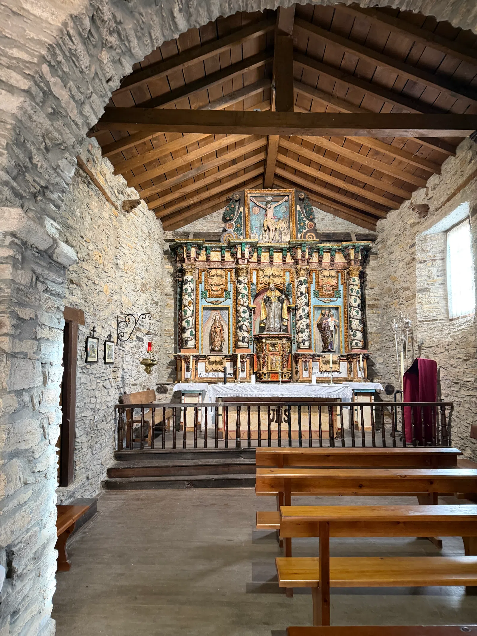 Interior of a small historic stone chapel with wooden ceiling, altar, religious statues, and wooden pews.