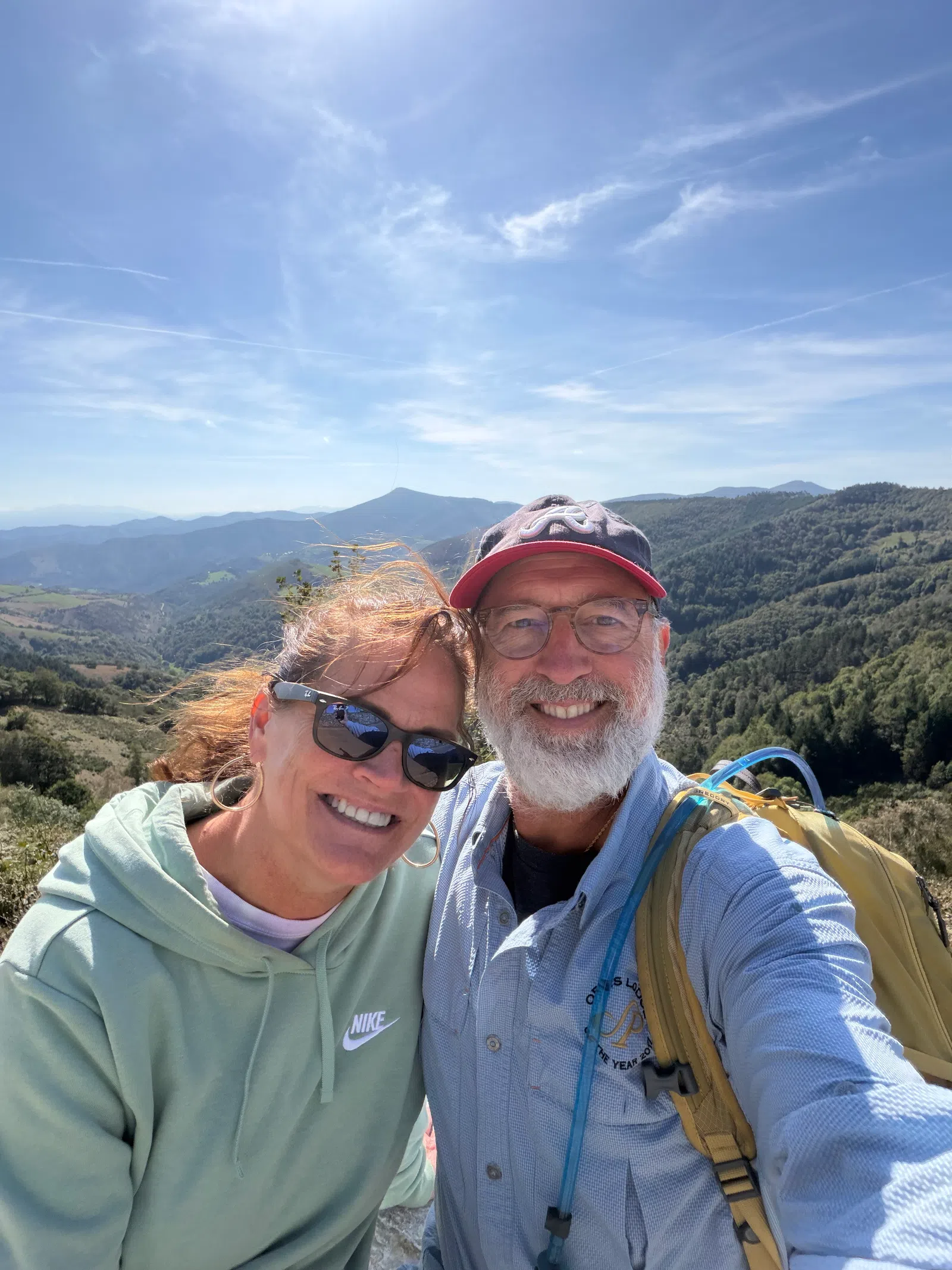 Smiling couple wearing sunglasses and hiking gear takes a selfie with mountain landscape under clear blue sky