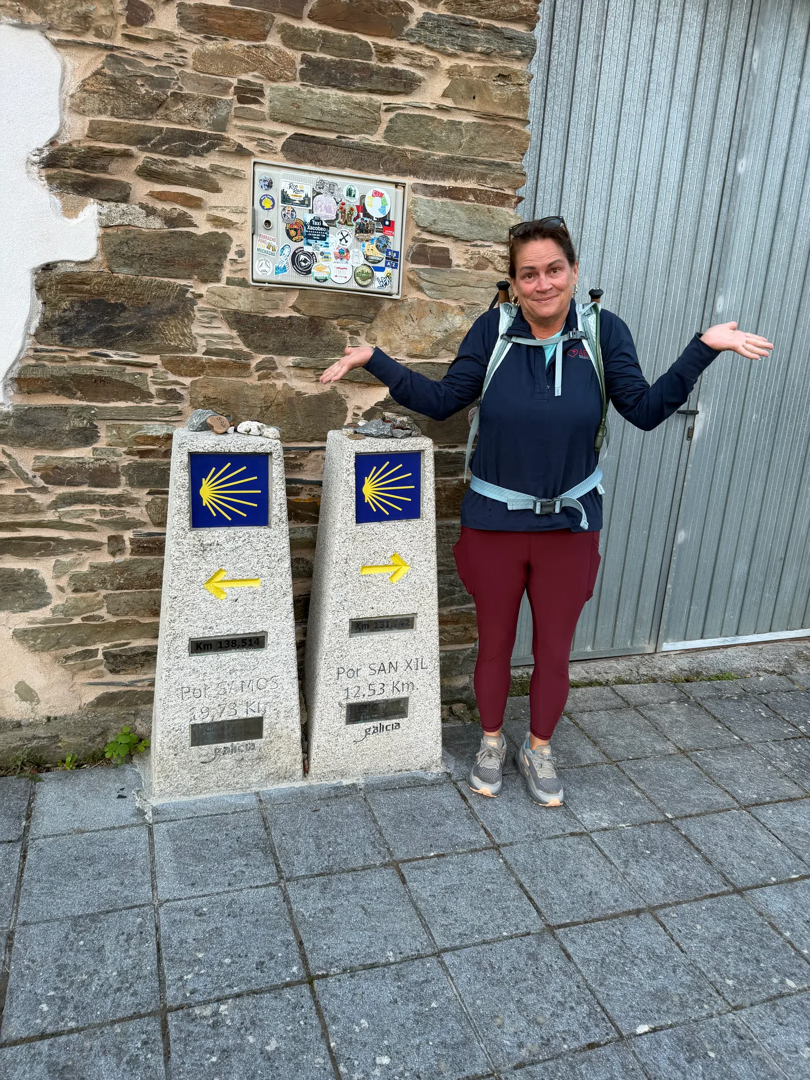 Woman with backpack stands near Camino de Santiago milestone markers showing different directions in Galicia, Spain.