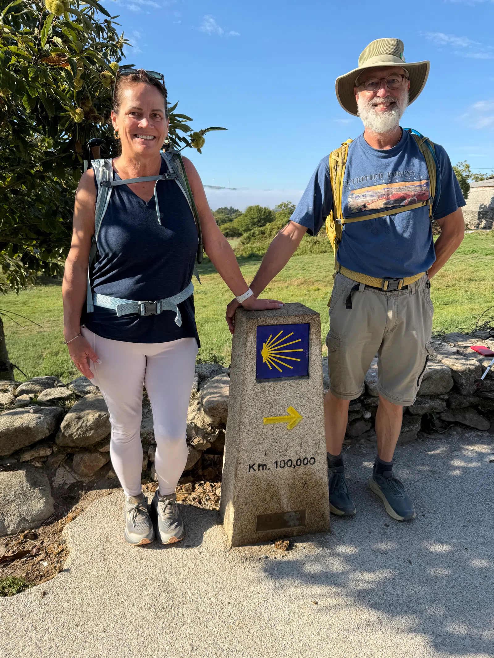 Two hikers standing beside a Camino de Santiago milestone marked 100,000 kilometers on a sunny day.