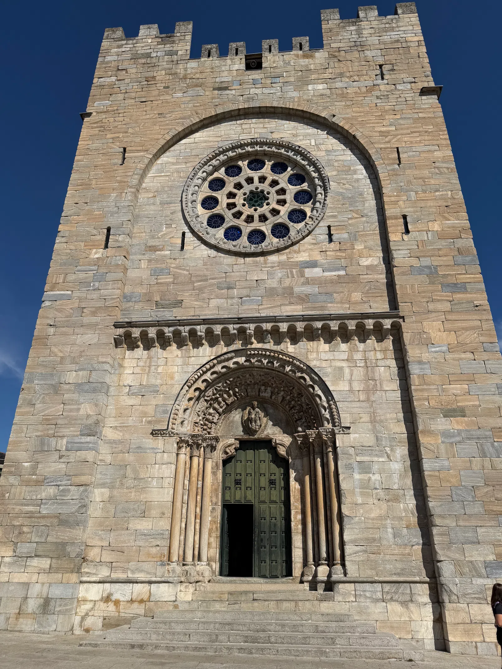 Stone cathedral tower with ornate arched doorway and large circular stained glass window under clear blue sky